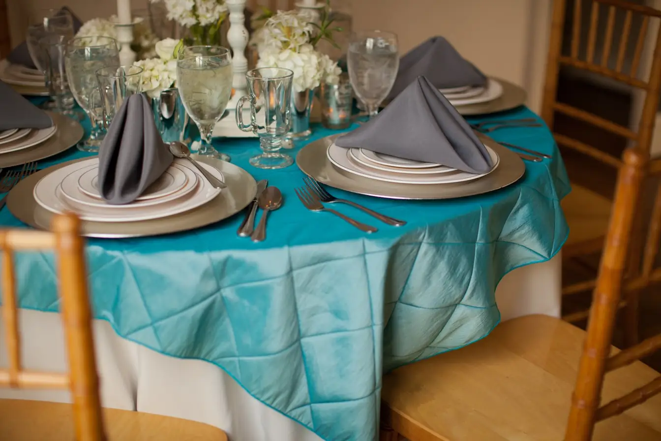 Silver-band white china on silver chargers with grey napkins and a teal pintuck overlay, fruitwood Chiavari chairs — china rentals from Party Connection Memphis
