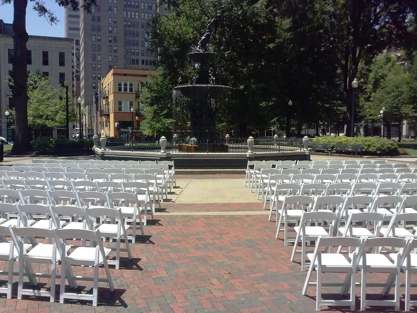 Rows of white folding chairs facing a tiered cast-iron fountain in a tree-lined downtown square for an outdoor ceremony