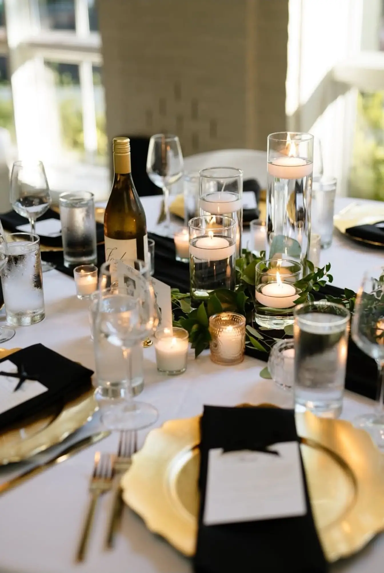Candlelit tablescape with floating candles in glass cylinders, greenery runner, gold scalloped chargers, and black napkins