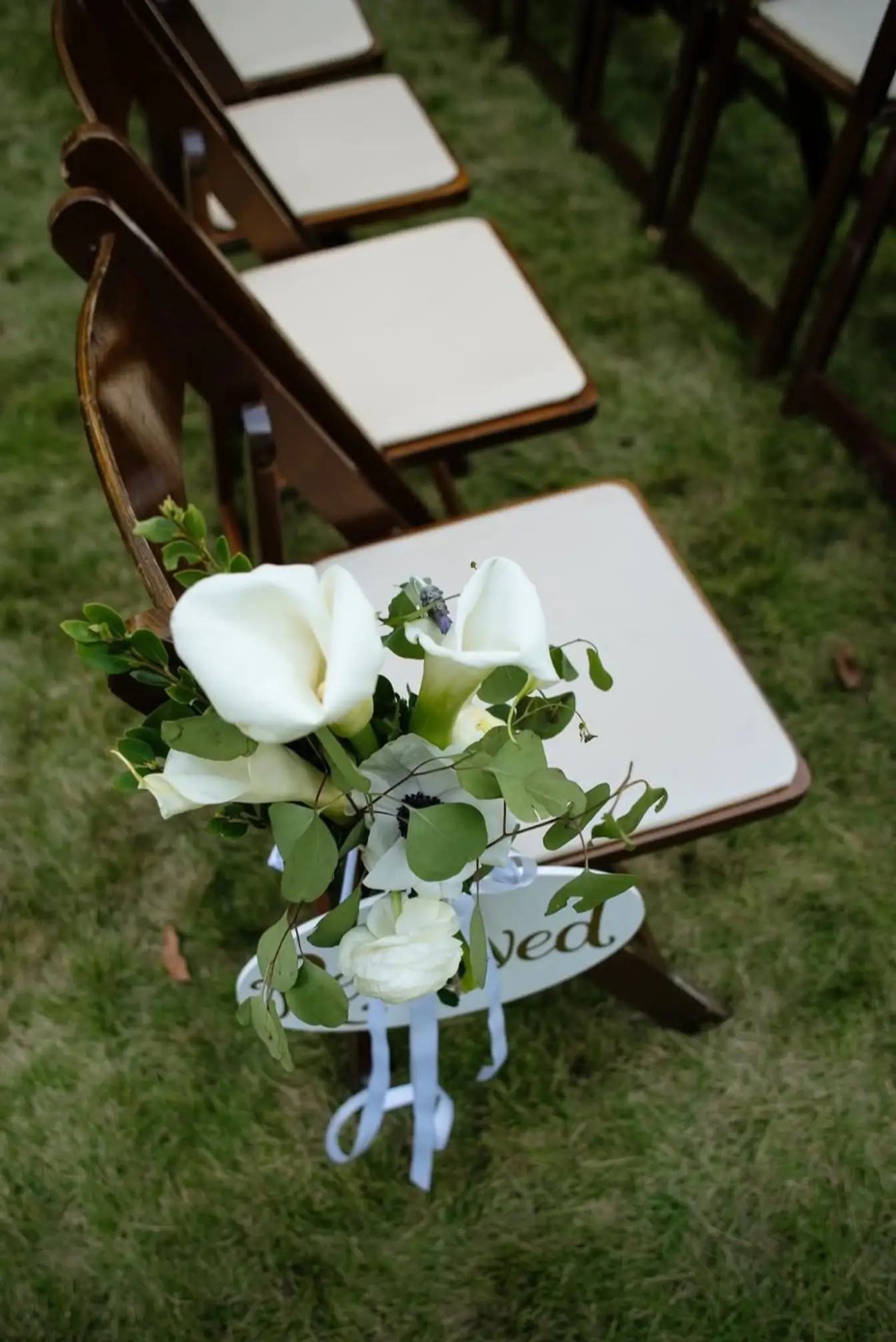 Calla lily and eucalyptus posy with a script sign hung on a dark wood folding chair along a ceremony aisle