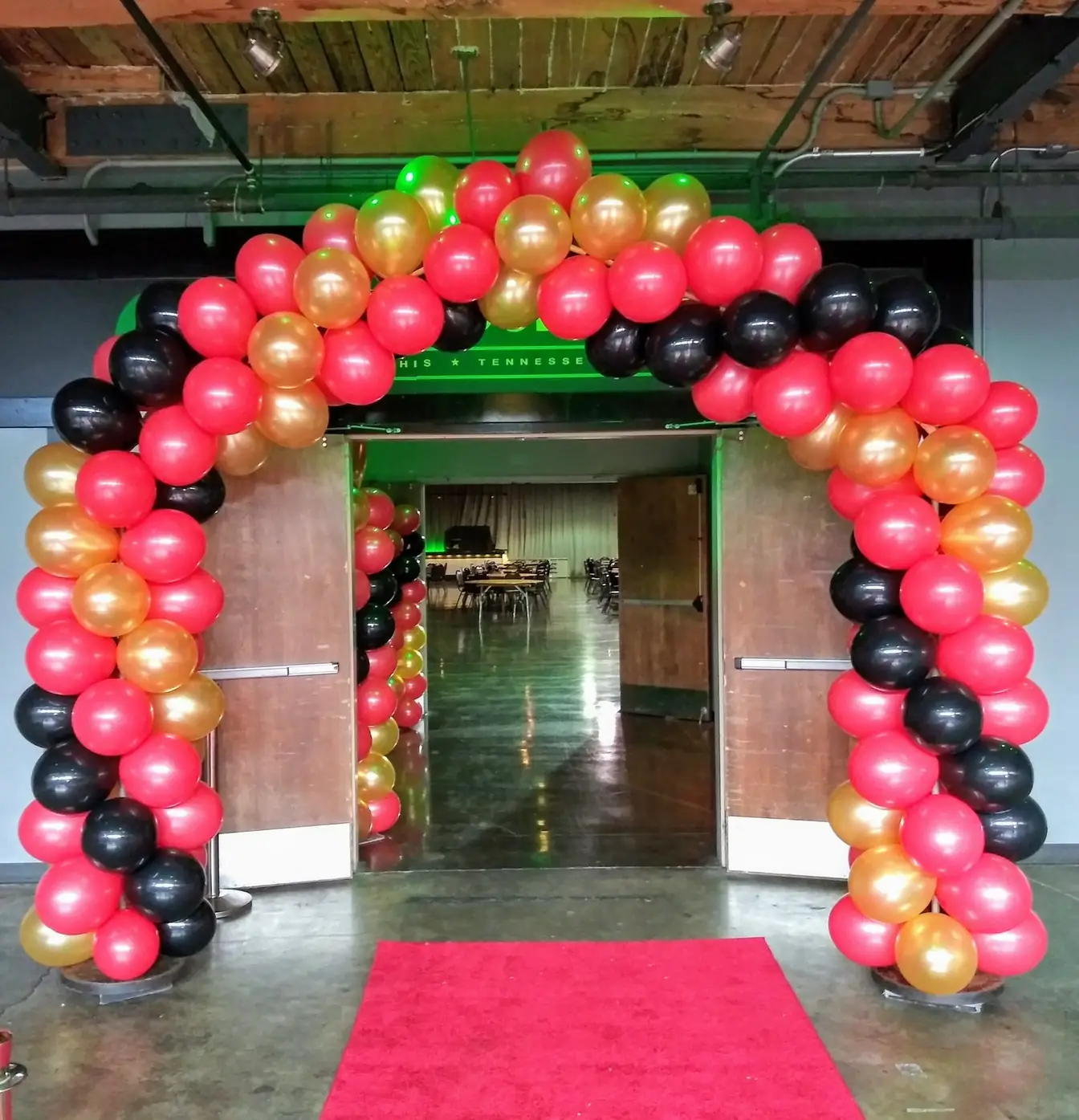 Red, black, and gold balloon arch framing open double doors with a red carpet laid beneath