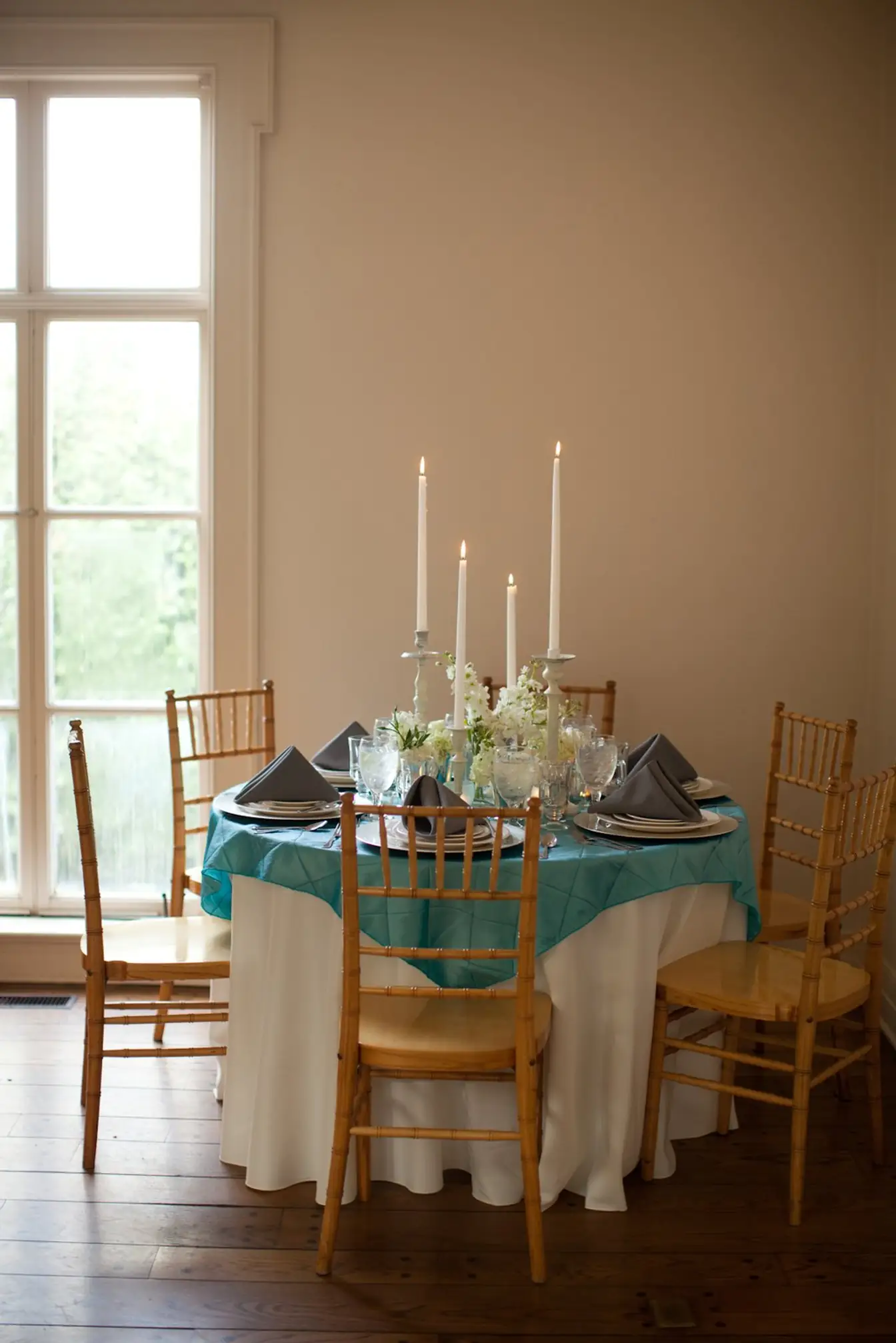 Round table in white floor-length linen with a teal pintuck overlay, gray napkins, and white taper candles beside a tall paned window