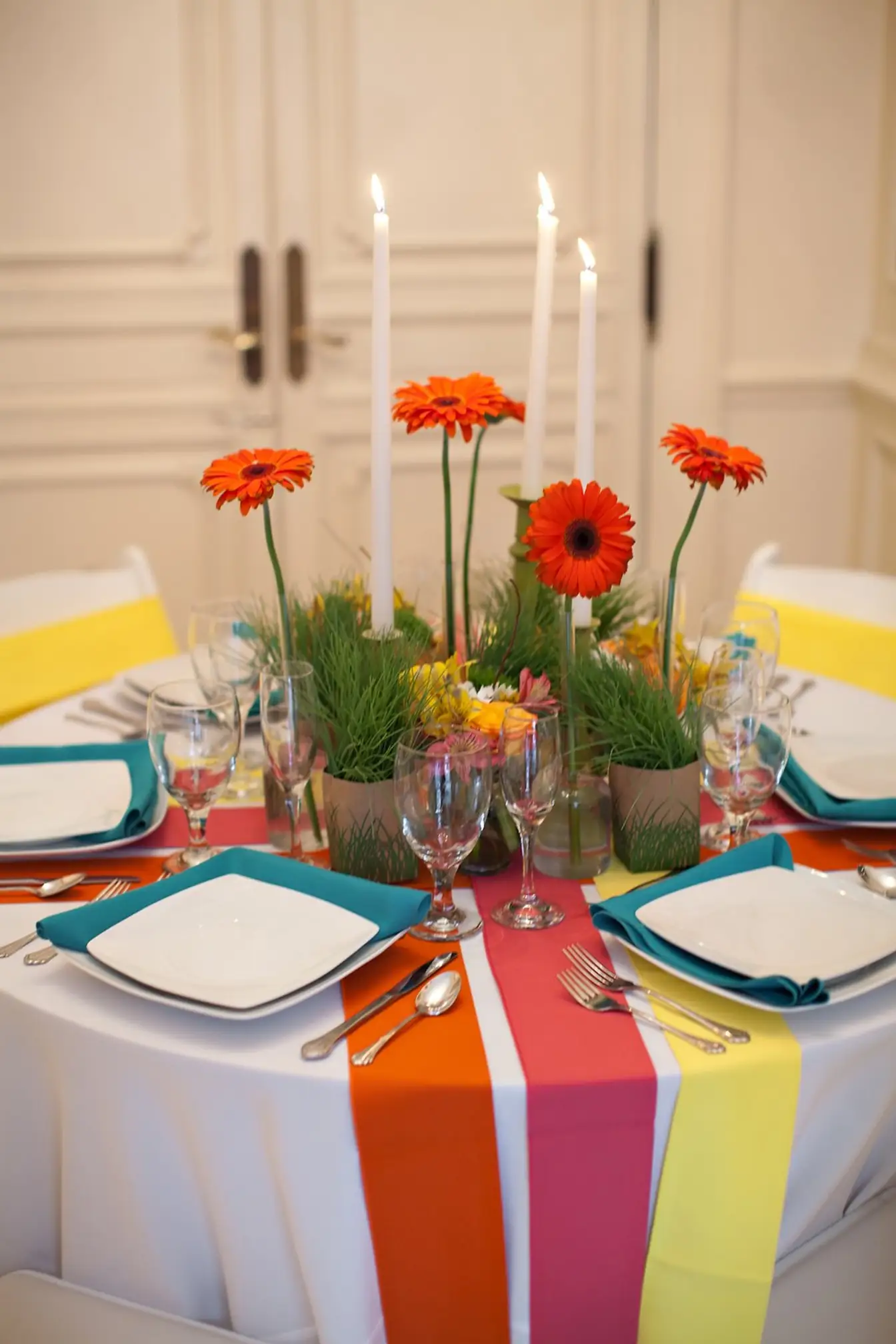 White round table crossed with orange, pink, and yellow runners, teal napkins on square plates, and orange gerbera daisy centerpieces