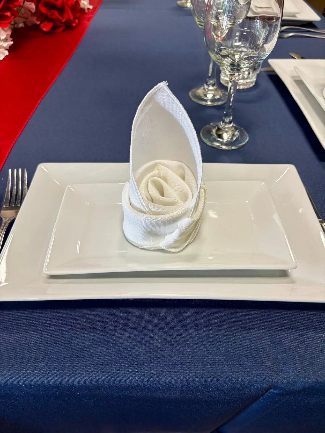 White napkin folded into a rose on stacked square white plates over navy linen with a red satin runner