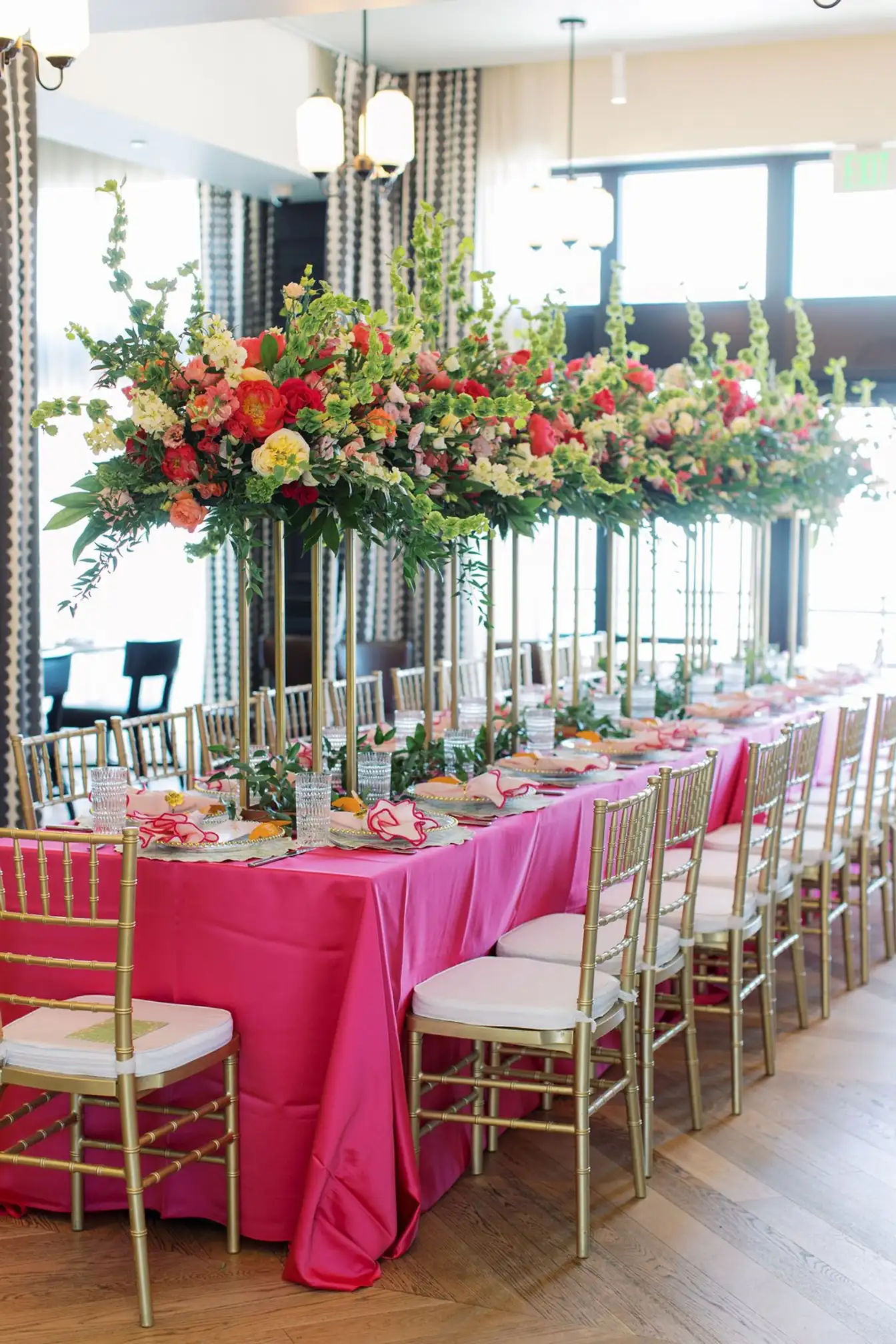 Long fuchsia satin banquet table with gold Chiavari chairs beneath elevated runners of coral, pink, and green florals on gold stands