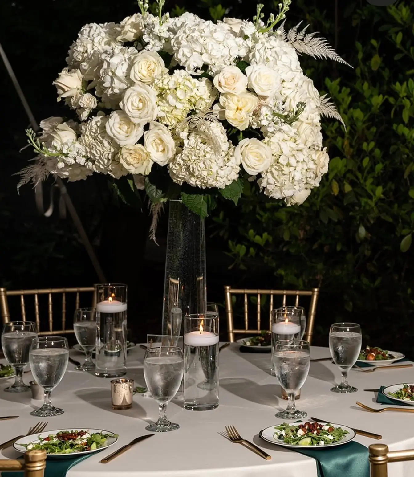 Tall centerpiece of white roses and hydrangeas above floating candles on a candlelit table set with salads and emerald napkins