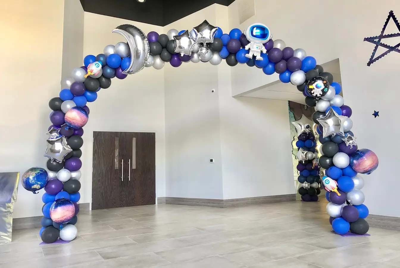 Space-themed balloon arch in navy, purple, silver, and black with astronaut, planet, moon, and star balloons spanning a lobby