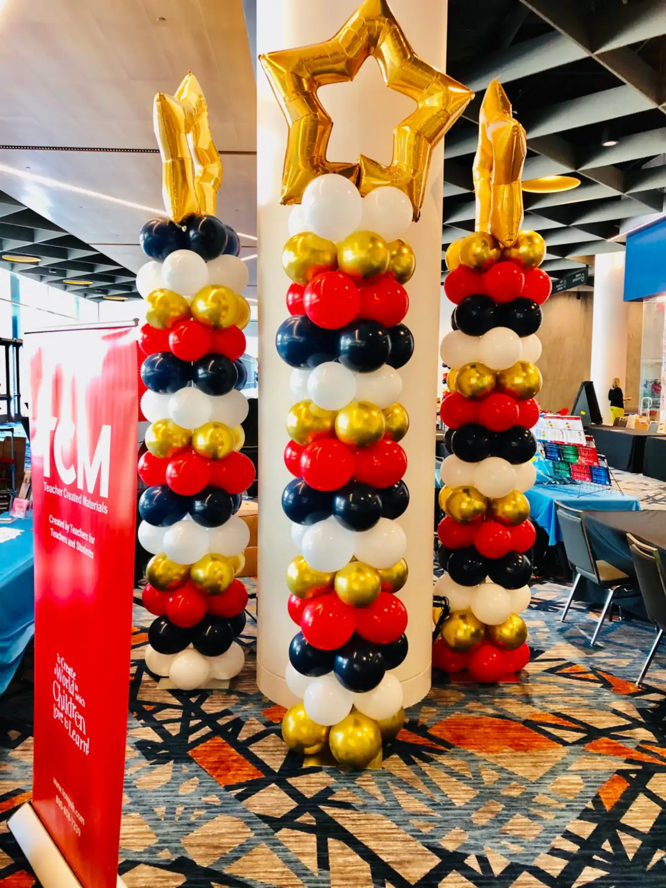 Three balloon columns in red, white, navy, and gold, each topped with a gold foil star, standing in a convention lobby