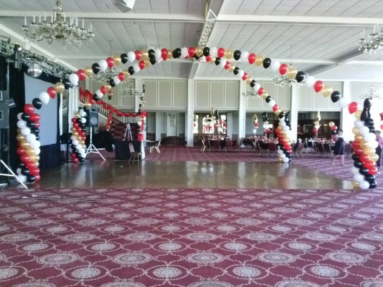 Red, black, gold, and white balloon arch spanning a ballroom dance floor, with spiral balloon columns at each end