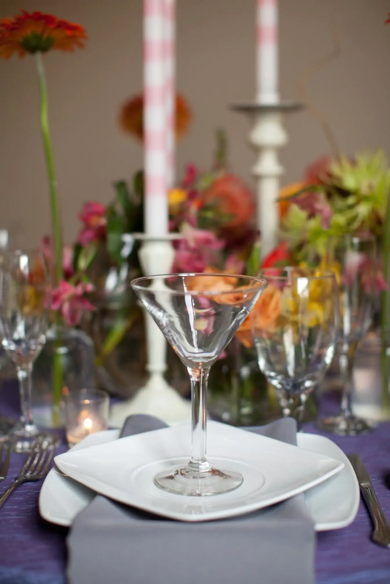Martini glass on stacked white square plates with a gray napkin over purple linen, gerbera daisies and pink-striped taper candles behind