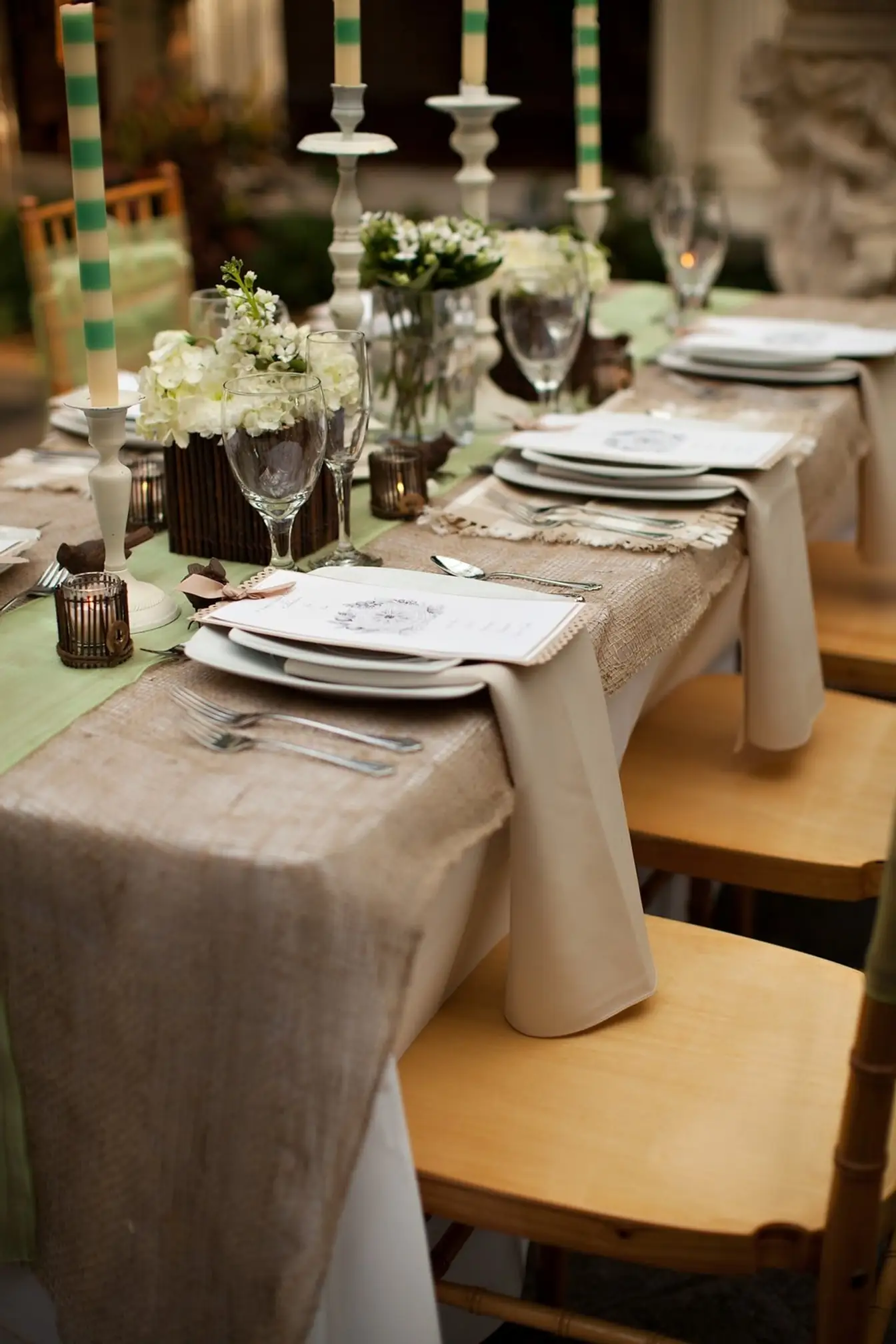 Long table with burlap and green runners over ivory linen, ivory napkins, white china with printed menus, striped taper candles, and gold Chiavari chairs
