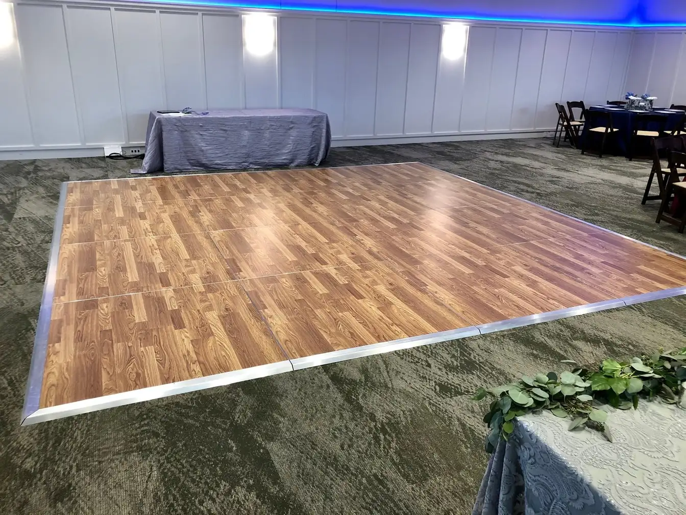 Portable wood-look dance floor laid on ballroom carpet beside a navy-linen round table with dark wood folding chairs and a crinkle-draped banquet table