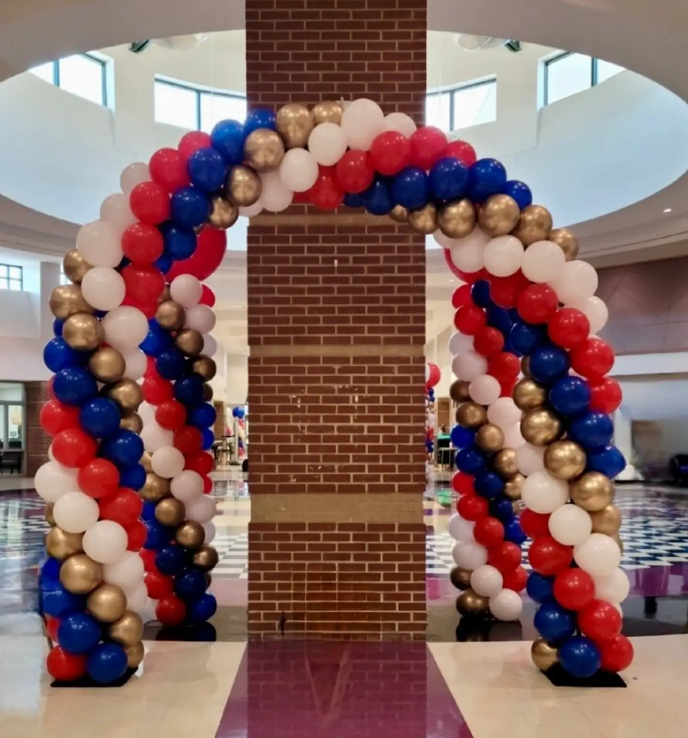 Red, white, royal blue, and gold balloon arch wrapping a brick column in a school lobby