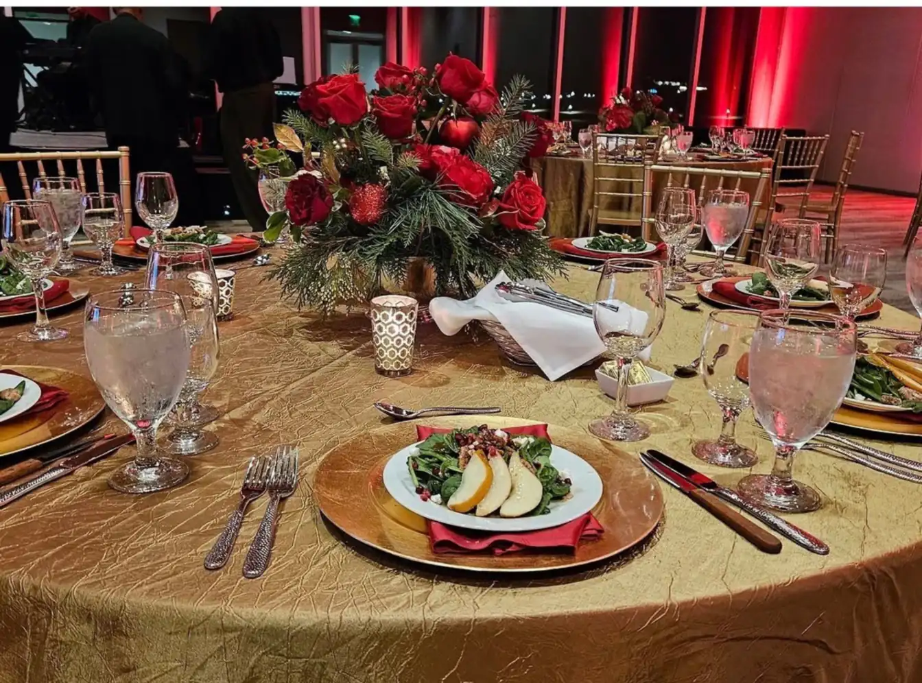 Gold crinkle linen set with gold chargers, white china, red napkins, hammered flatware, and red roses, ringed by gold Chiavari chairs under red uplighting