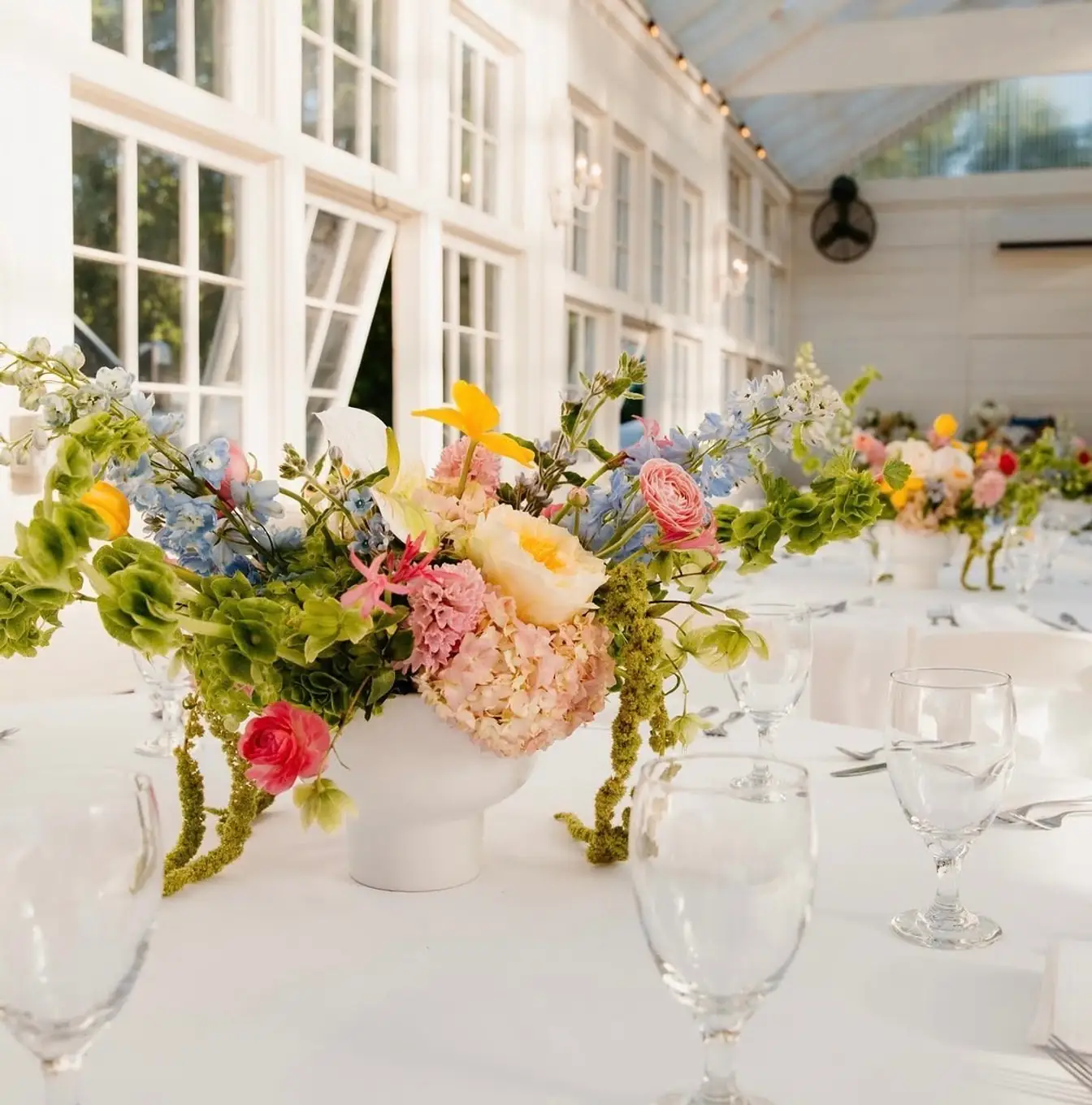 White linen with water goblets and flatware and a low centerpiece of pink hydrangea, blue delphinium, and yellow blooms in a sunlit white conservatory