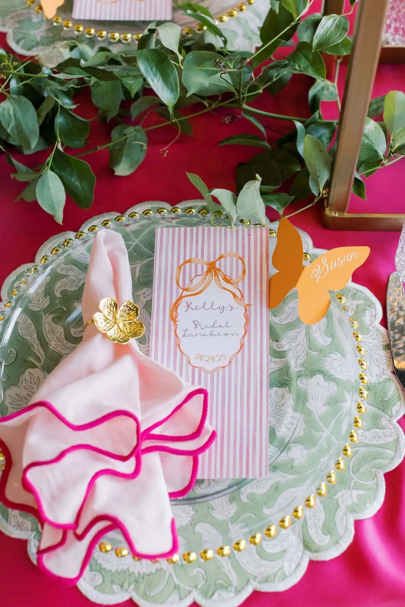 Gold-beaded charger with a green patterned plate, pink scalloped napkin in a gold flower ring, and a striped menu card on hot pink linen with greenery