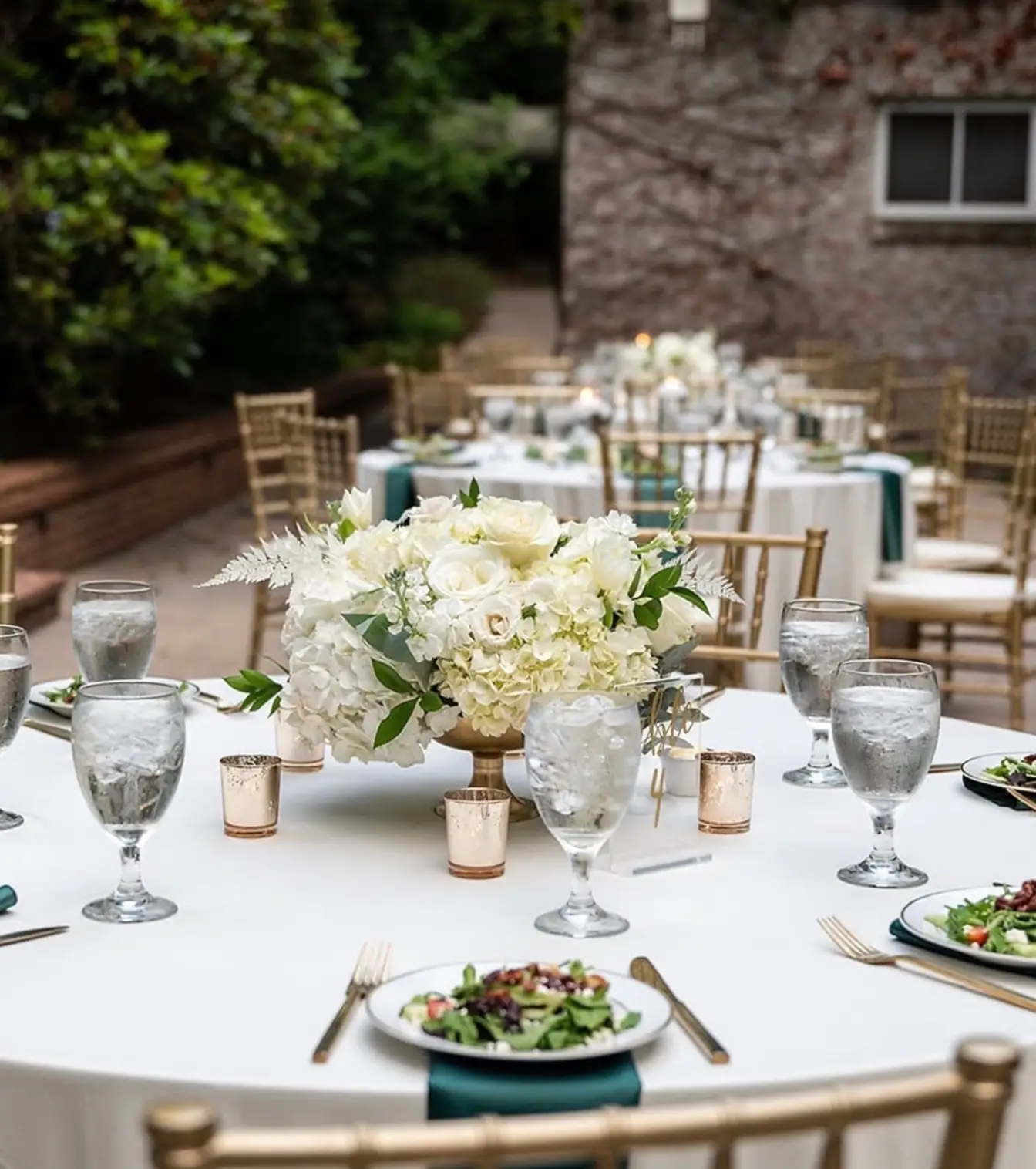 Courtyard reception table in white linen with emerald napkins, gold flatware, a white hydrangea and rose centerpiece, and gold Chiavari chairs