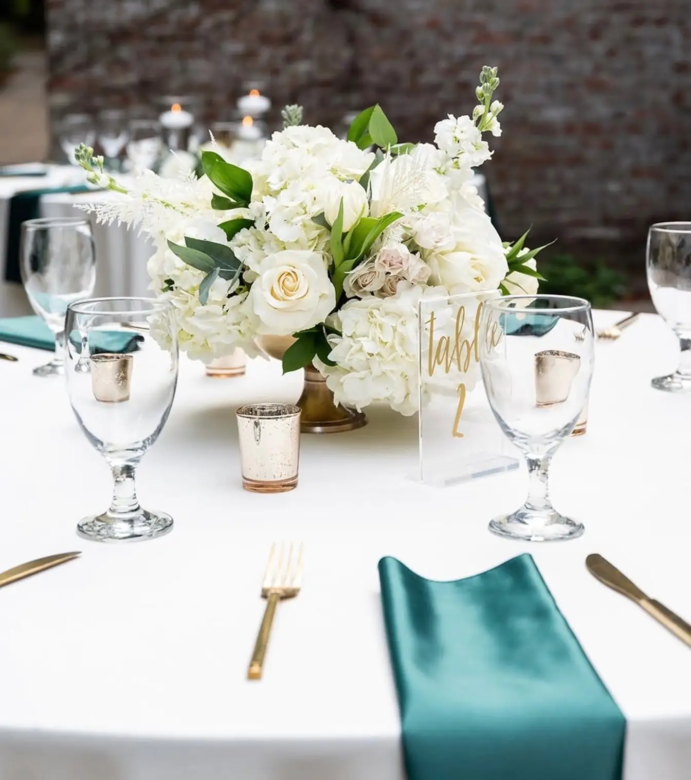 Close-up of a white linen table with an emerald satin napkin, gold flatware, water goblets, mercury-glass votives, and a white rose centerpiece
