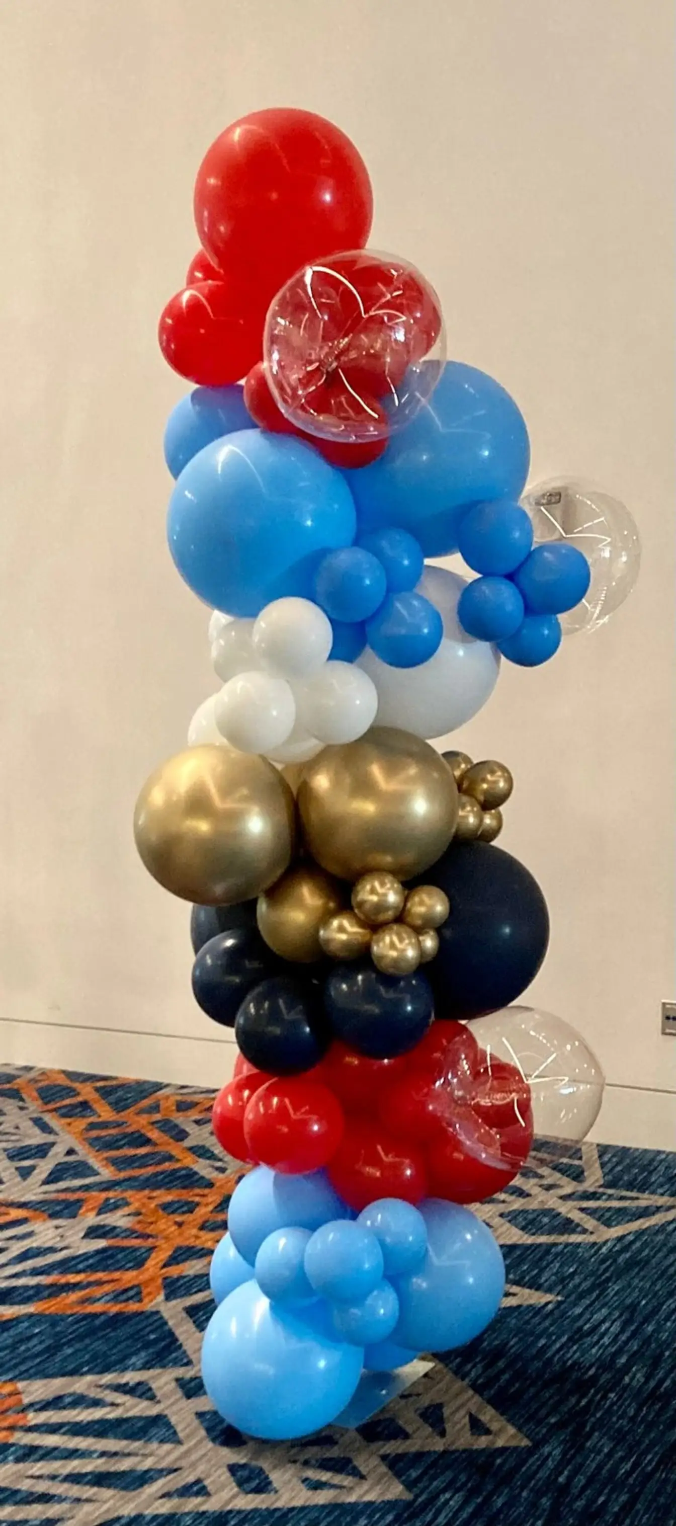 Organic balloon column in red, light blue, white, gold, and navy with clear confetti balloons in a convention hall