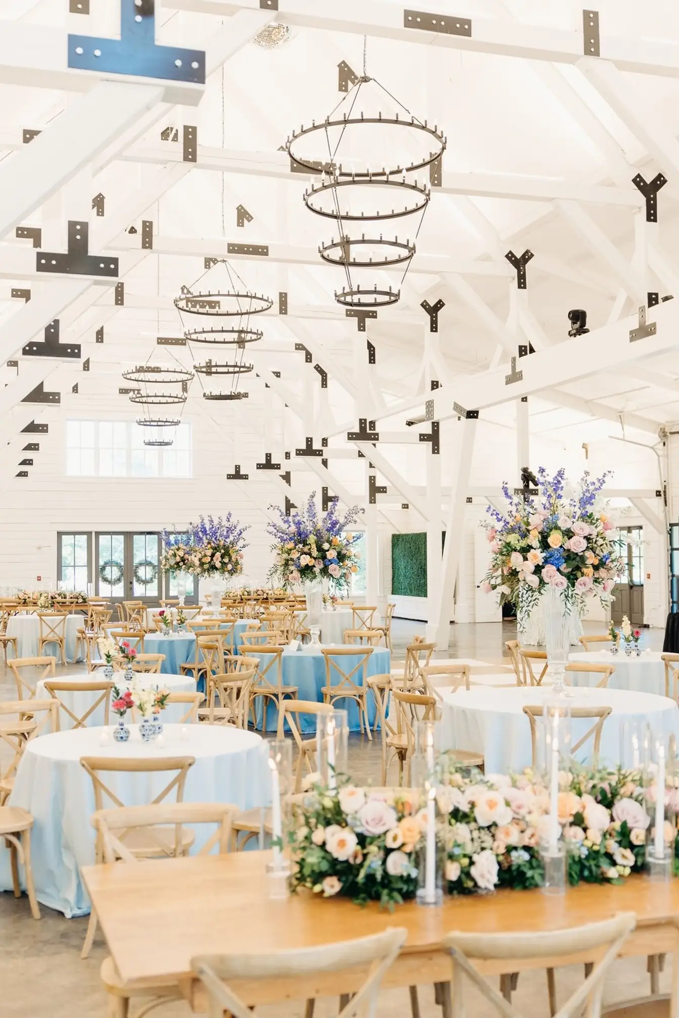 White-beamed barn reception with round tables in light blue linens, tall pastel florals, iron ring chandeliers, and a candlelit farm head table