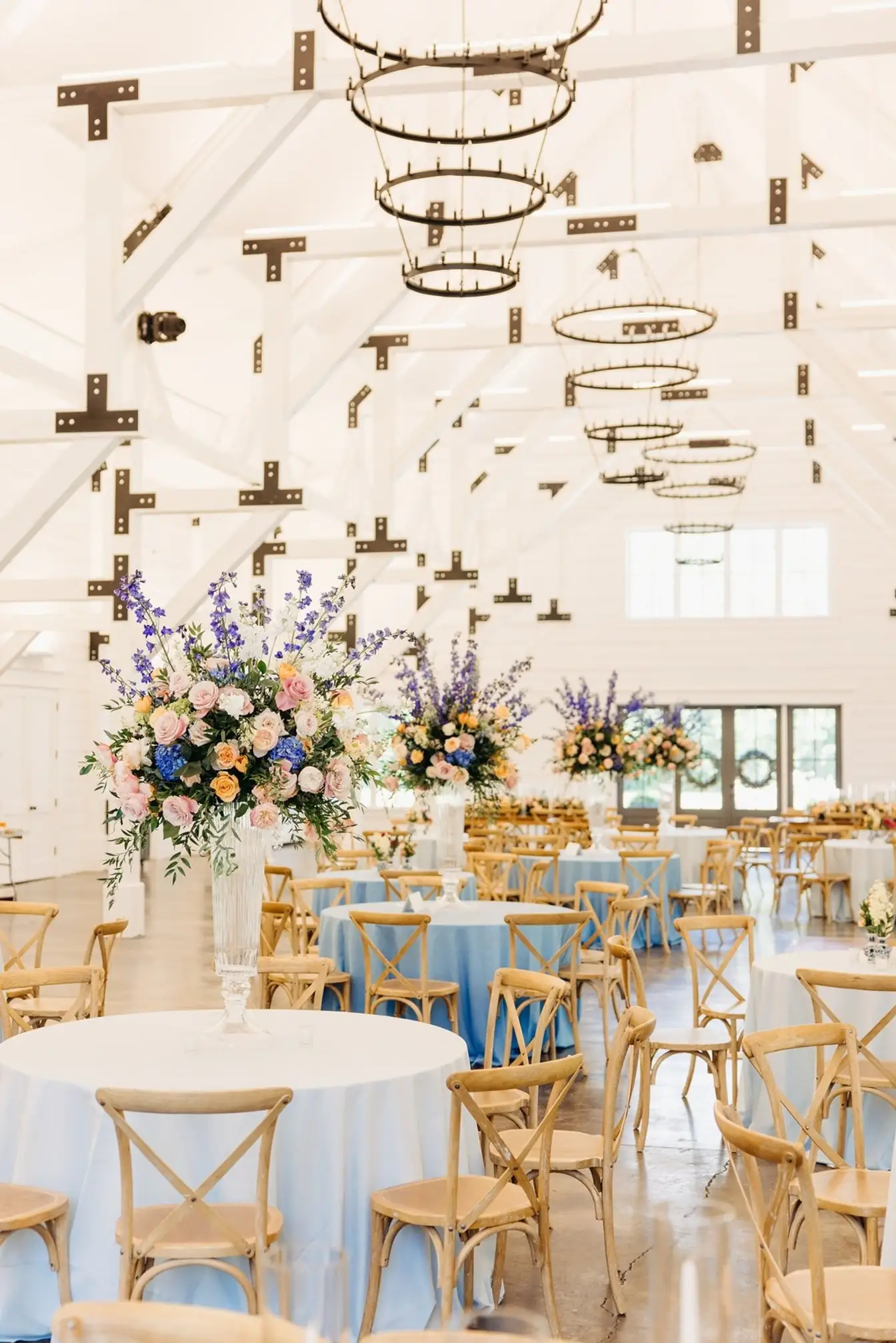 Round tables in light blue linens with towering pink, peach, and blue floral arrangements in a bright white barn