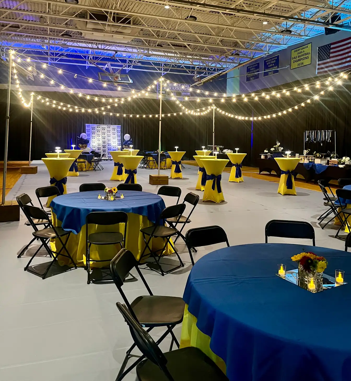 Royal blue round tablecloths with yellow and black chairs under bistro lights in a school gym