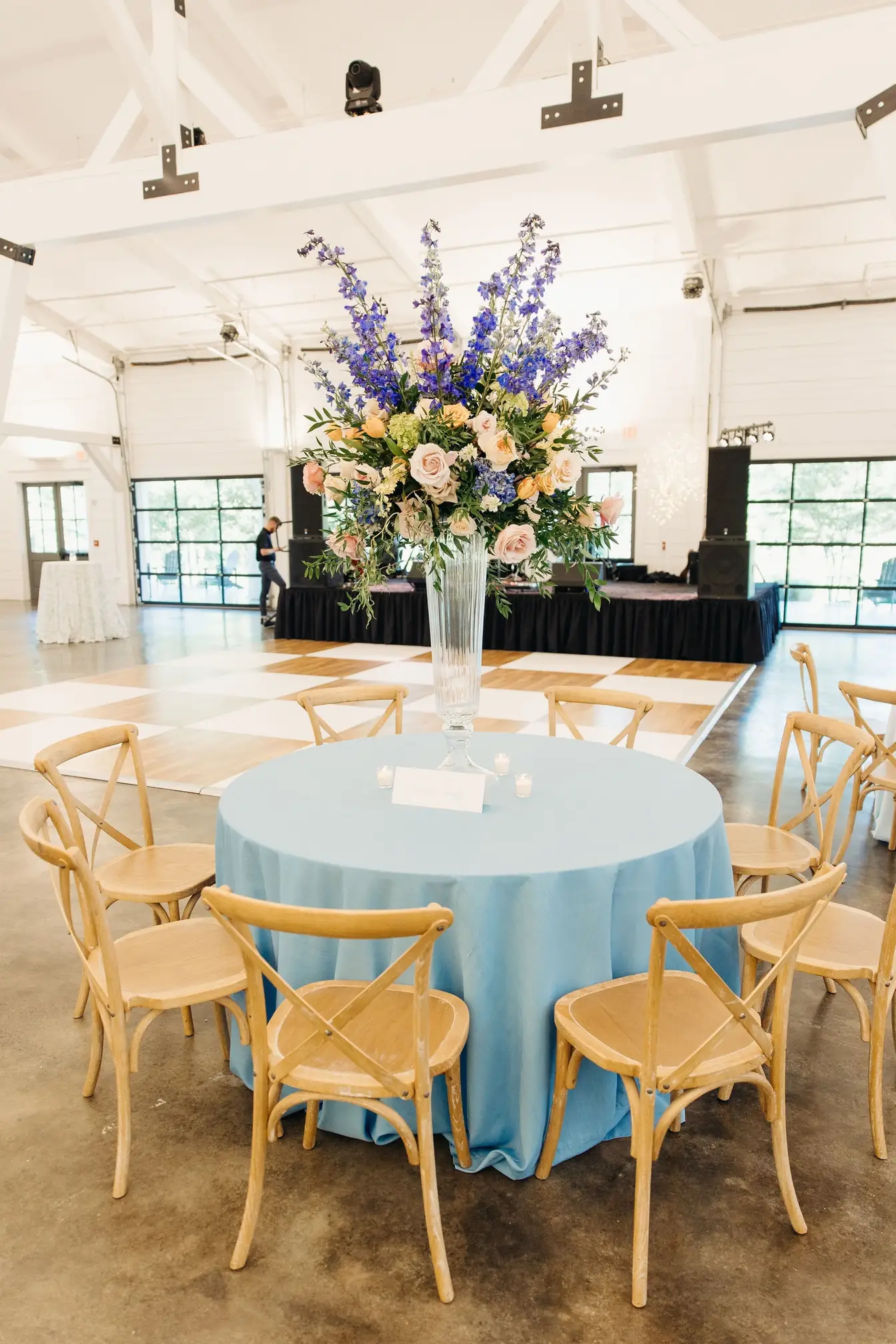 Round table in light blue linen with a tall pastel floral centerpiece and cross-back wood chairs in a white barn