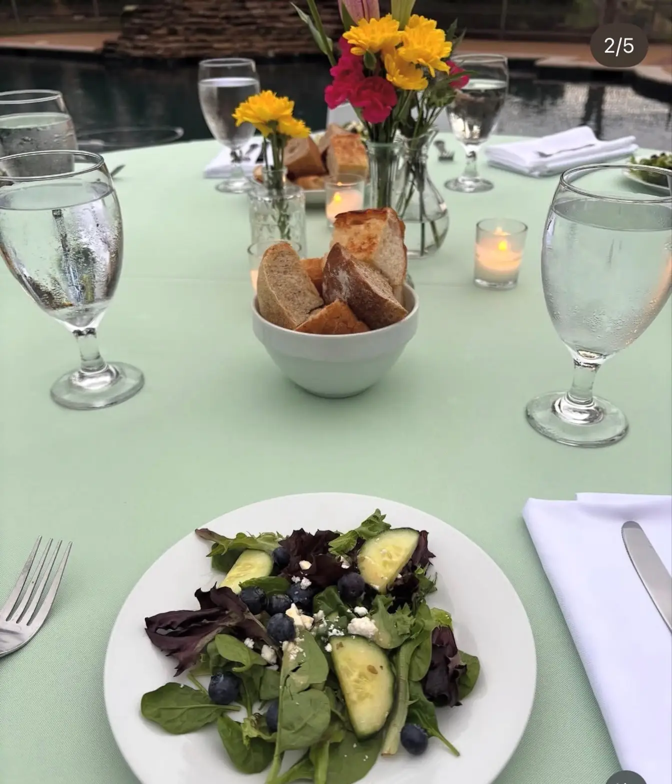 Sage green tablecloth set with a plated salad, a china bread bowl and water goblets at a poolside dinner