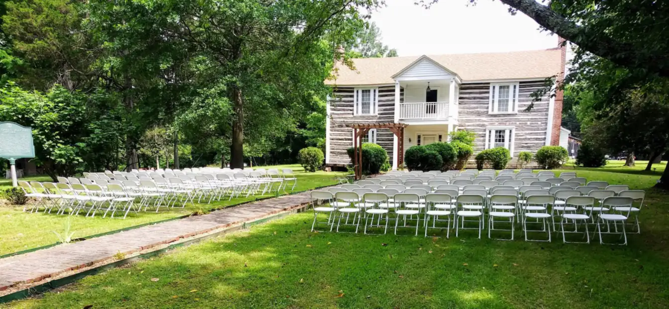 Rows of white folding chairs arranged on the lawn of a historic two-story log home for an outdoor ceremony, framed by large trees