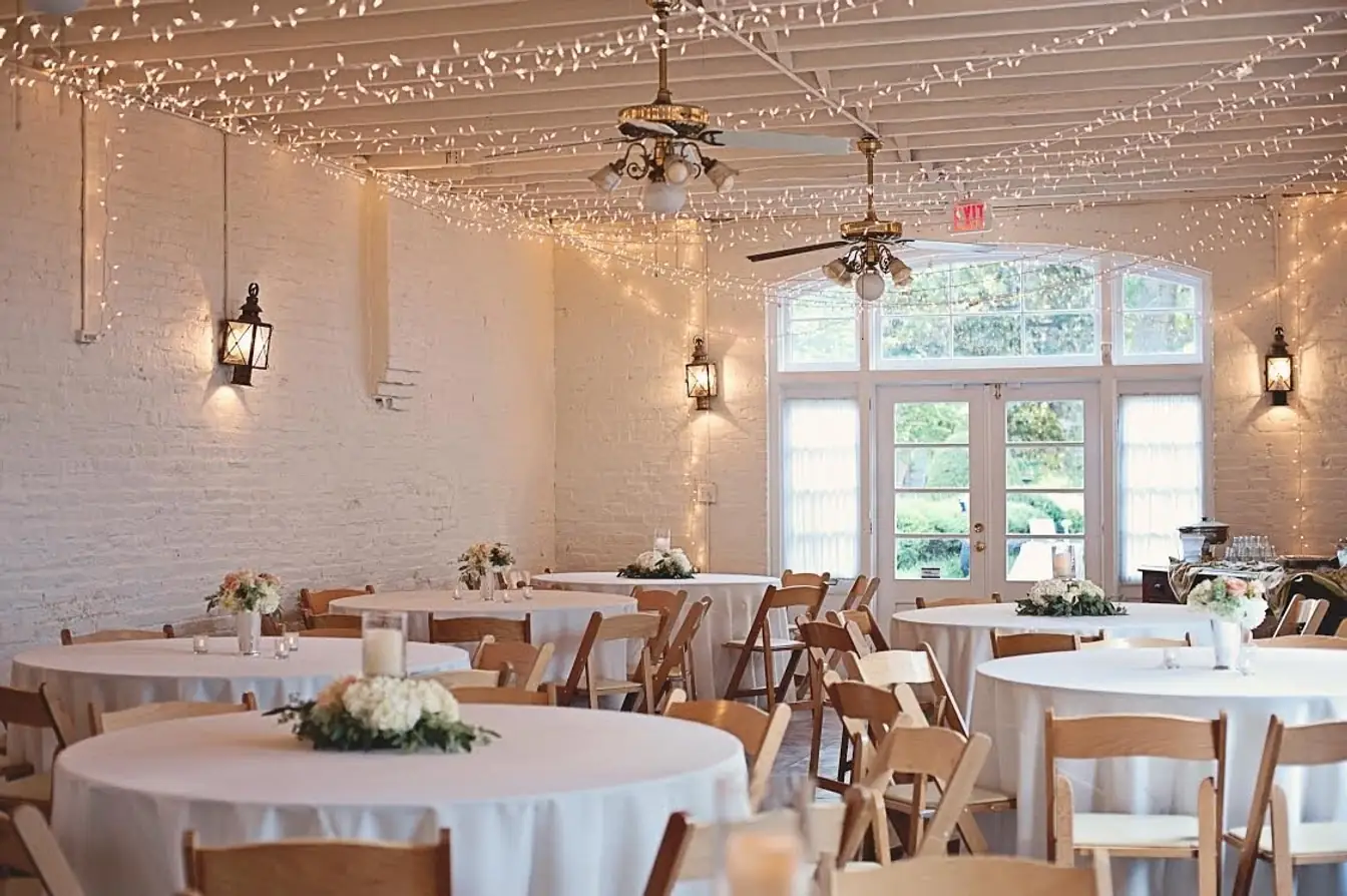 White-brick hall strung with fairy lights — round tables in floor-length white linens with fruitwood folding chairs and small rose centerpieces