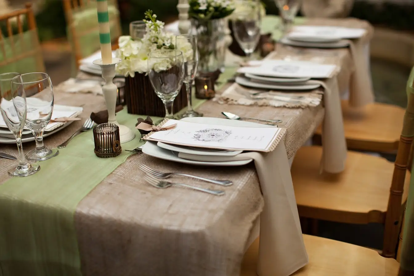 Head table styled with burlap and sage green runners, stacked ivory china with printed menu cards, glassware, and gold Chiavari chairs