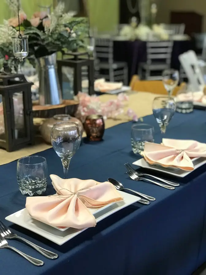 Head table in navy linen and burlap runner set with blush pinwheel-folded napkins on square white plates, with lanterns and soft florals down the center