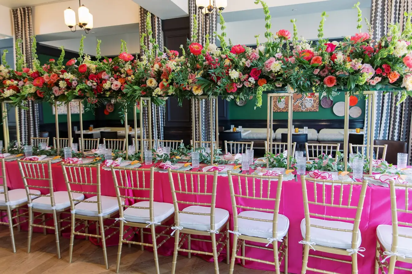 Long hot-pink satin banquet table lined with gold Chiavari chairs beneath elevated arrangements of coral peonies, roses, and greenery