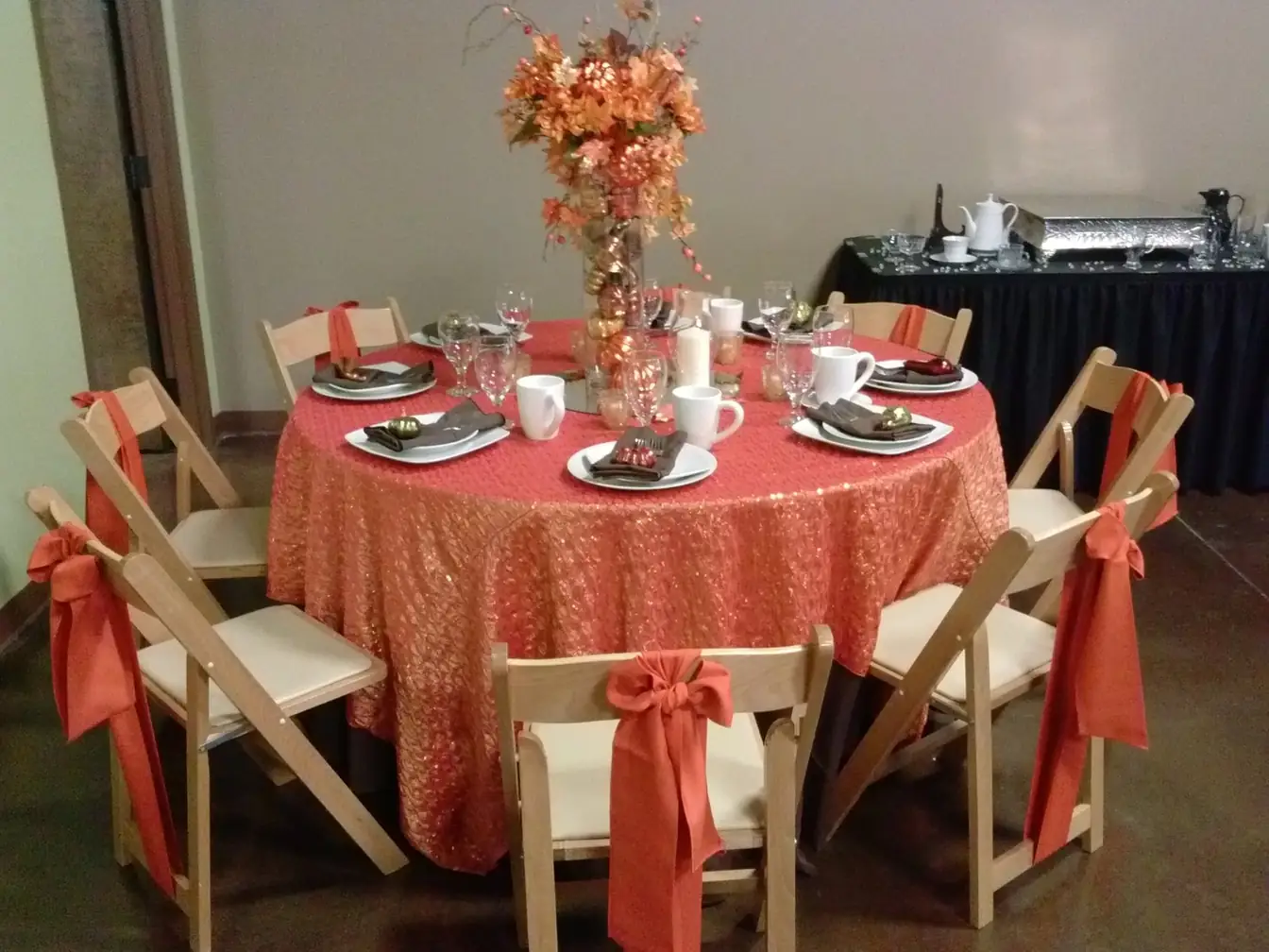 Round table in a coral sequin overlay with a fall branch centerpiece and place settings of mugs and cocoa, ringed by natural-wood folding chairs sashed in burnt orange