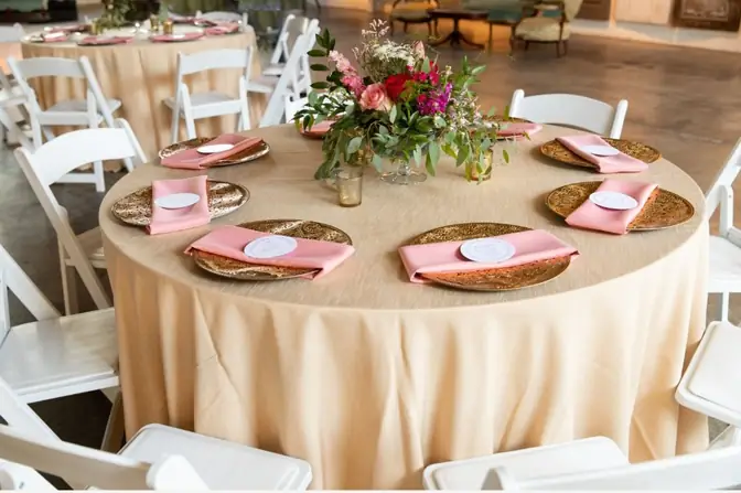 Round table in champagne linen set with gold charger plates, pink napkins, and a rose and wildflower centerpiece, ringed by white padded folding chairs