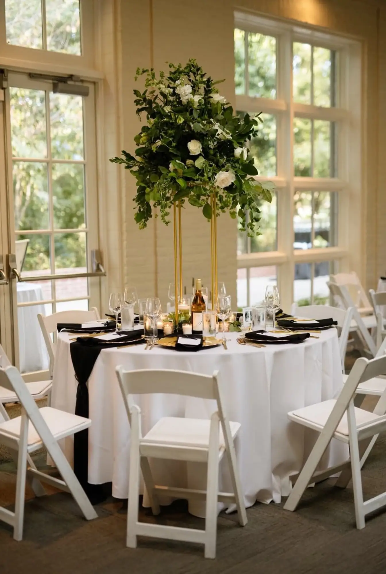 Round table in white floor-length linen with black napkins, gold-rimmed chargers, and a tall greenery-and-white-rose centerpiece in a sunlit garden room