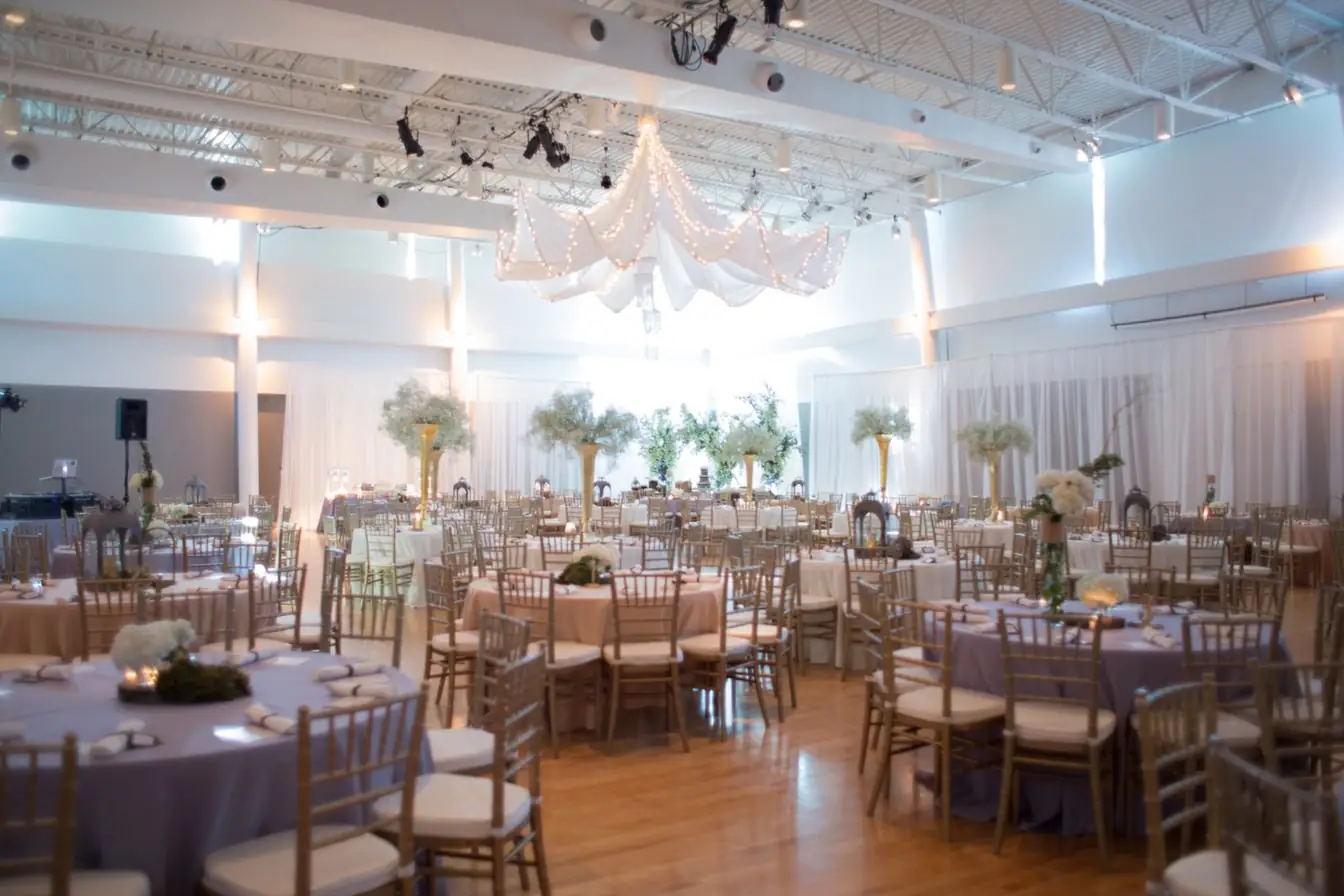 Ballroom of round tables in lavender and blush linens with gold Chiavari chairs, tall gold trumpet centerpieces, and a lighted fabric canopy overhead