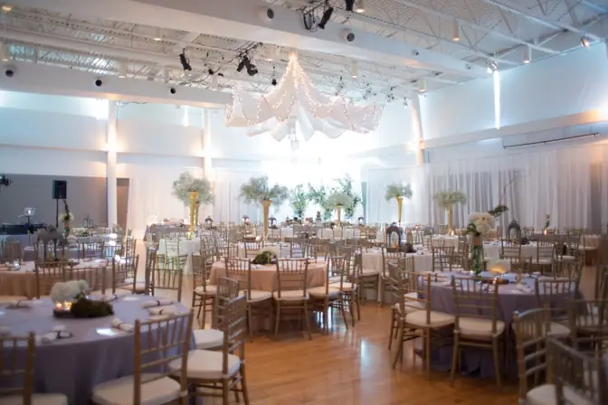 Ballroom of round tables in lavender and blush linens with gold Chiavari chairs, tall gold trumpet centerpieces, and a lighted fabric canopy overhead