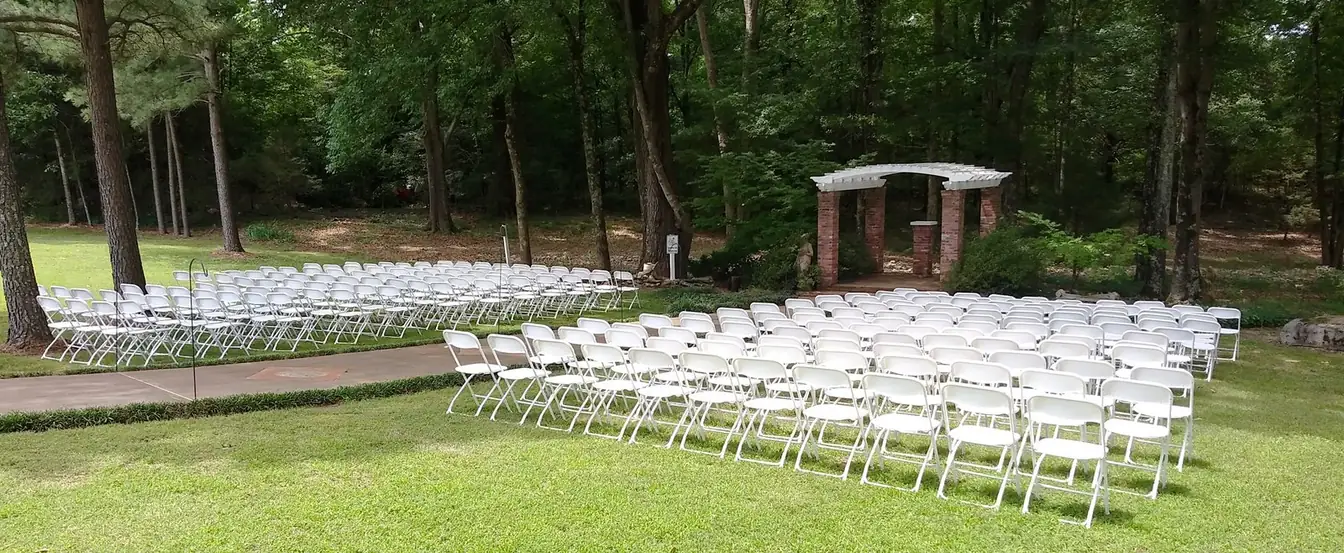 White folding chairs fanned across a shaded lawn facing a brick-and-timber pergola for a wooded outdoor ceremony