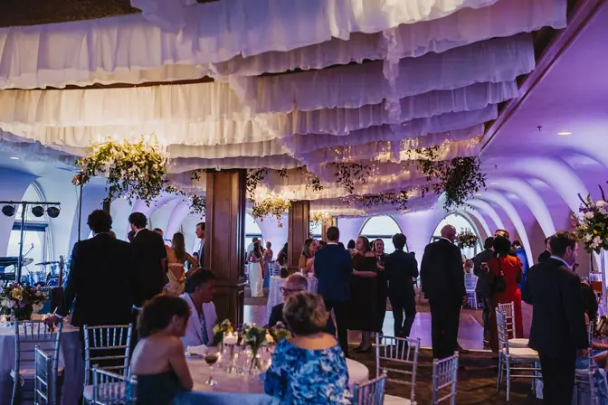 Wedding reception beneath white ruffled ceiling draping and hanging greenery, with guests mingling around tables set with silver Chiavari chairs