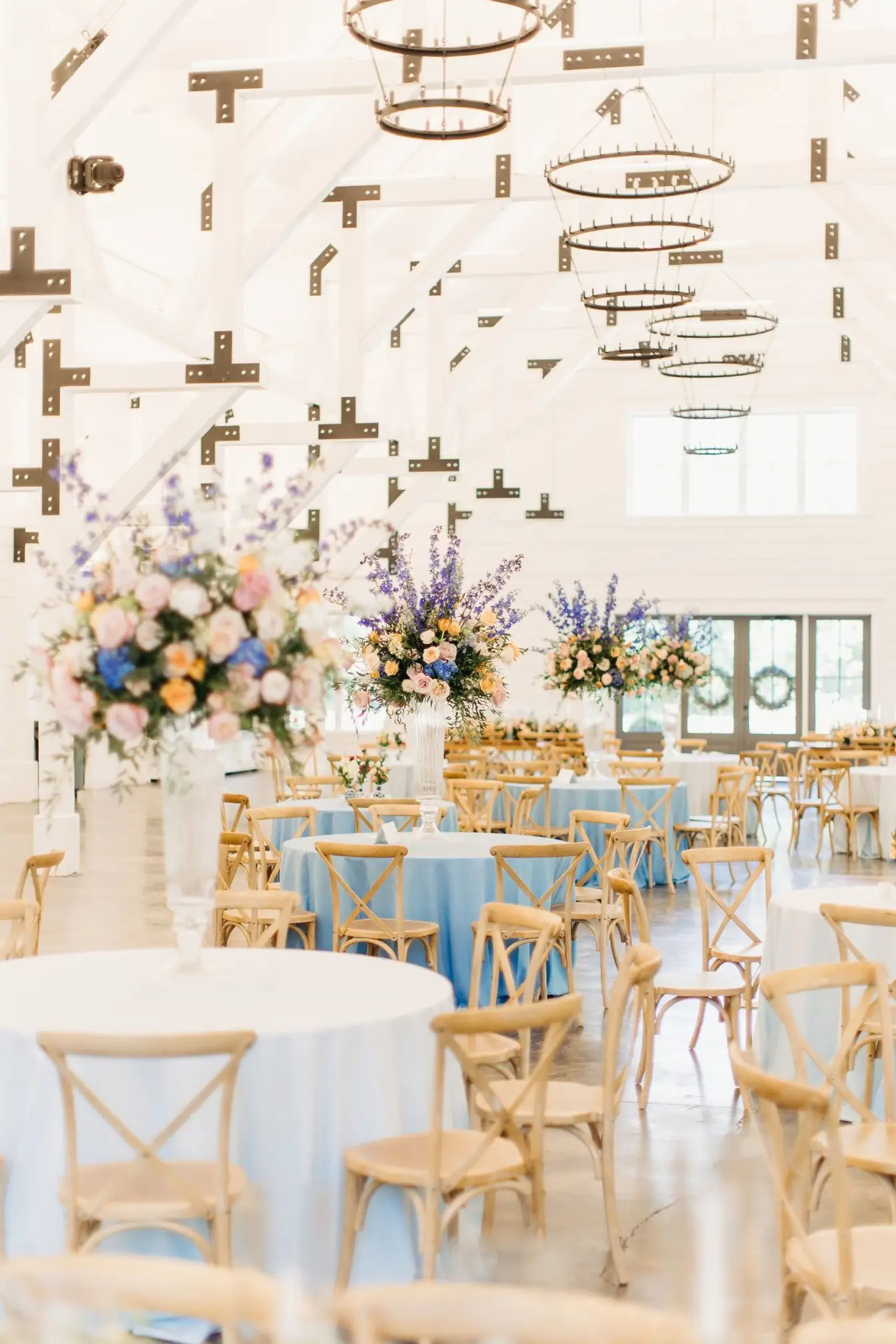 Bright white-beamed venue hall filled with round tables in pale blue linens, wooden cross-back chairs, and tall pastel floral arrangements