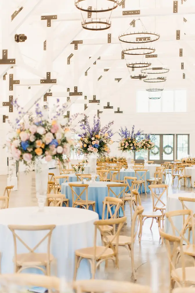 Bright white-beamed venue hall filled with round tables in pale blue linens, wooden cross-back chairs, and tall pastel floral arrangements
