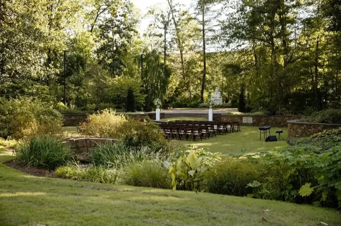 Rows of dark wood folding chairs arranged on a lawn for a garden ceremony, framed by landscaped beds, ornamental grasses, and tall trees