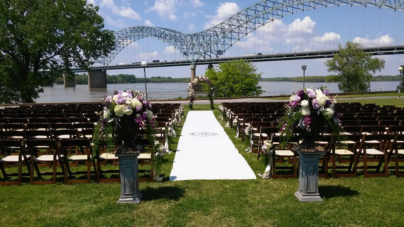 Outdoor wedding ceremony with rows of dark wood folding chairs, purple and white floral urns on pedestals, and a white monogrammed aisle runner on a riverside lawn beneath a steel arch bridge