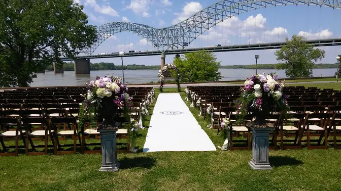 Outdoor wedding ceremony with rows of dark wood folding chairs, purple and white floral urns on pedestals, and a white monogrammed aisle runner on a riverside lawn beneath a steel arch bridge