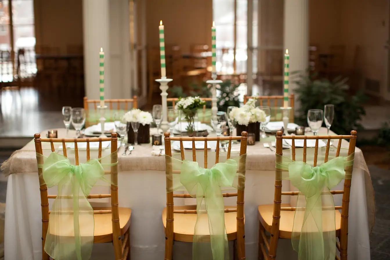 Table with burlap runner, green-striped taper candles, and white flower posies, seated with gold Chiavari chairs tied in green organza sashes