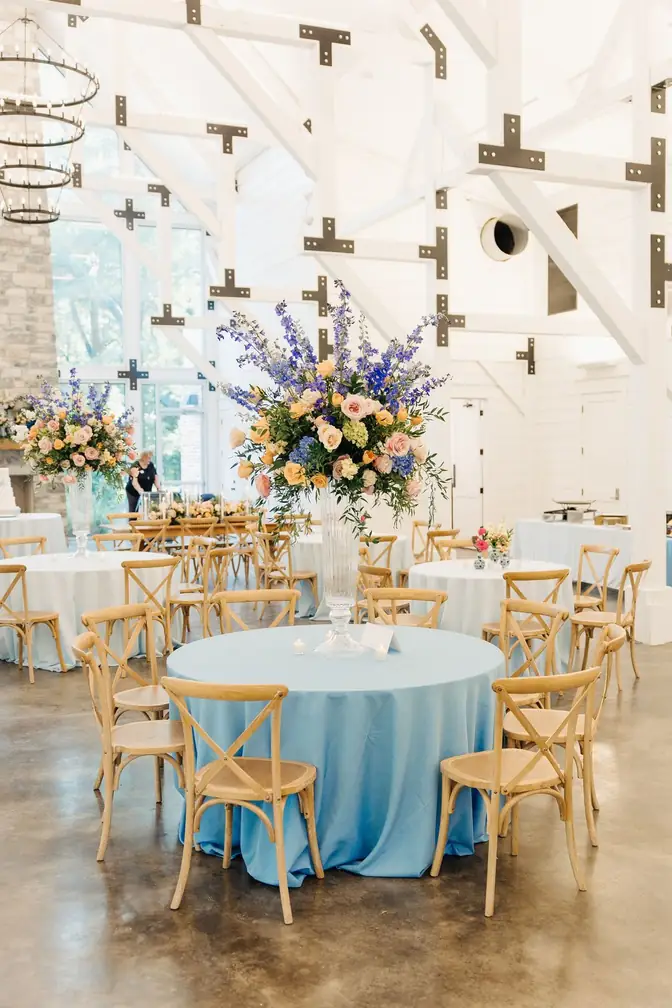 Round table in cornflower-blue linen ringed by wooden cross-back chairs, topped with a towering centerpiece of delphinium, roses, and peach blooms