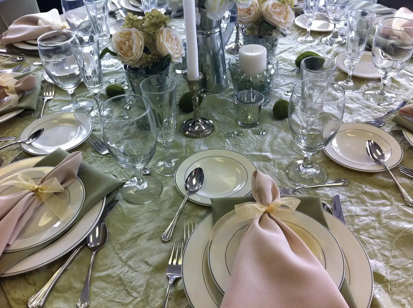 Table set in sage crinkle linen with ivory china, blush napkins tied in sheer gold ribbon, silver candlesticks, and ivory rose centerpieces