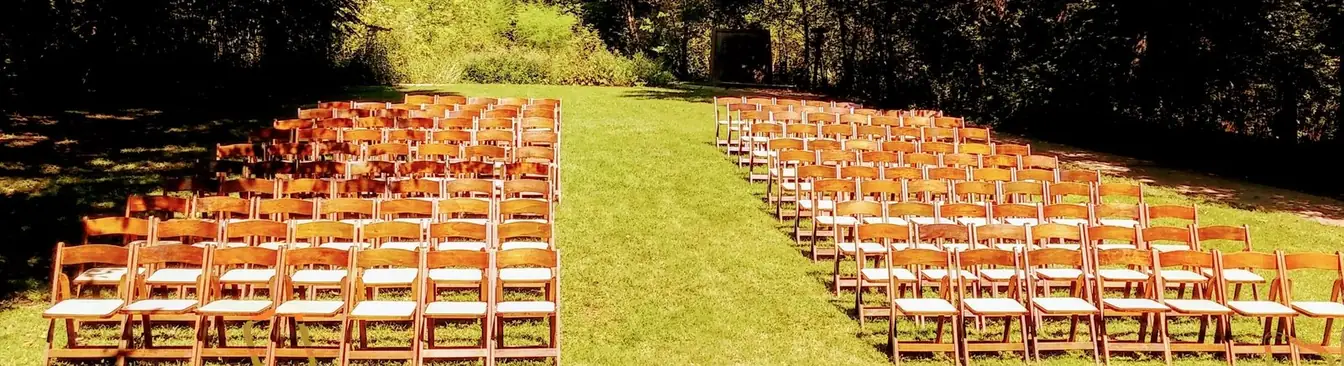 Rows of wooden folding chairs with padded seats arranged on a sunlit lawn with a wide grass aisle between the sections