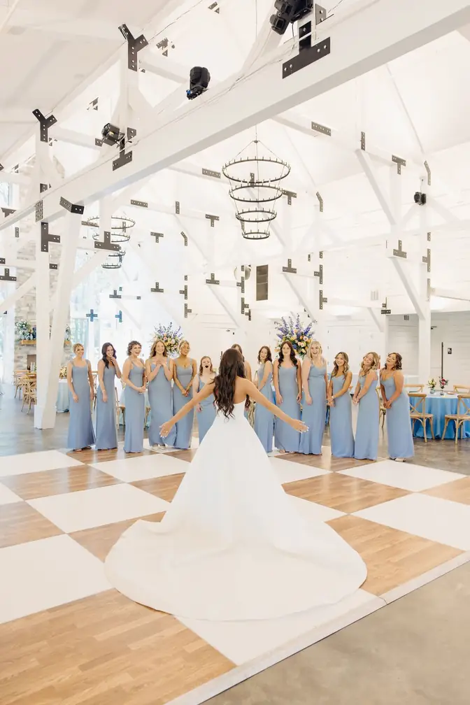Bride in a full white gown standing on a white-and-wood checkered dance floor with arms open toward her bridesmaids in dusty-blue dresses